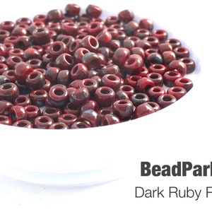 May include: A close-up of a bowl filled with dark ruby red Picasso seed beads. The beads are small and round, with a slightly iridescent finish.