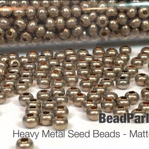 May include: A close-up of a pile of small, round, matte gold metal seed beads. The beads are in a clear container and are scattered on a white surface. The text "BeadParlorBeads Heavy Metal Seed Beads - Matte 24K Gold" is visible at the bottom of the image.