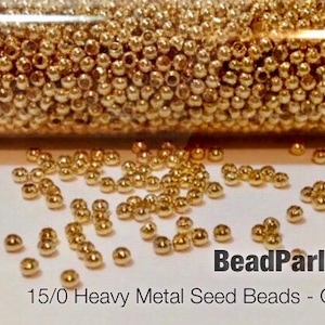 May include: A close-up of a container filled with small, gold-colored metal seed beads. The beads are scattered on a white surface. The text "BeadParlorBeads 15/0 Heavy Metal Seed Beads - Gilding Gold" is visible at the bottom of the image.