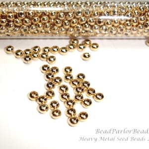 May include: A close-up of a container filled with small, shiny gold metal seed beads. The beads are scattered on a white surface. BeadParlor Beads Heavy Metal Seed Beads 24k Gold.