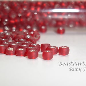May include: Close-up of a small glass tube filled with red glass beads. The beads have a frosted finish and a silver sheen. The text "BeadParlorBeads Ruby Frost" is visible below the beads.
