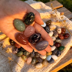 Op de afbeelding: Een verzameling gepolijste edelsteen eieren in verschillende kleuren, waaronder groen, roze, bruin en paars. De eieren worden in een hand gehouden en liggen op een crèmekleurig, getextureerd oppervlak, met een paar takjes gedroogde bloemen.