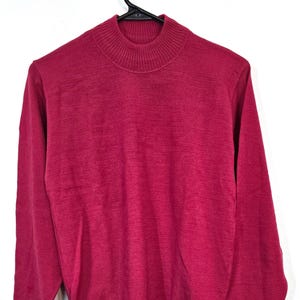 May include: A magenta-colored sweater with a mock neck and long sleeves hangs from a black hanger. The sweater has a ribbed neckline and cuffs. A tag with the brand name "Pendleton" is attached to the bottom of the sweater.