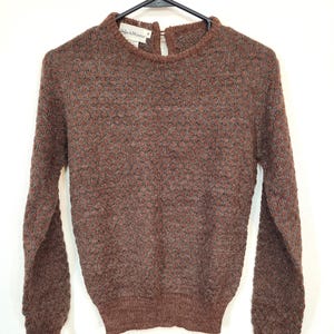 May include: A brown long-sleeved sweater with a textured pattern. The sweater has a high neckline and ribbed cuffs. The label reads "Black Winter". The sweater is hanging on a black hanger.