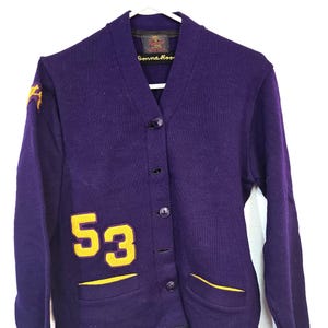 以下が含まれることがあります： 紫色のDonna Reedカーディガン。イエローのアクセントがあり、前面にイエローの数字「53」とポケットのトリムがあります。「Donna Reed」と書かれたラベルが付いています。