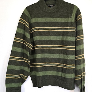 以下が含まれることがあります： 緑とベージュのストライプのセーター、モックタートルネック付き。セーターは長袖で、白い背景に黒いハンガーに掛けられています。「Towncraft」のブランド名が襟元に見えます。
