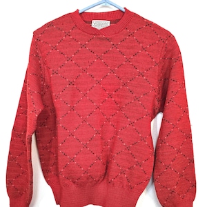 May include: A red argyle sweater with a crew neck and long sleeves. The sweater features a diamond pattern with small black and white accents. The sweater is made of a knit material and has ribbed cuffs and hem. A small tag is visible near the neckline.