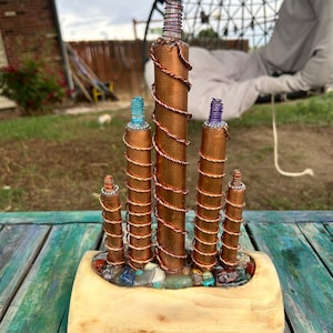 May include: A decorative sculpture featuring five copper-colored cylindrical pillars of varying heights, each wrapped in thin copper wire. The tops of the pillars are accented with colored wire coils, and they are set into a light-colored wooden base.