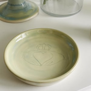 Può includere: Un piccolo piatto rotondo in ceramica verde pallido con un design Claddagh. Il simbolo Claddagh, con un cuore, una corona e mani, è in rilievo al centro. Il piatto ha un bordo leggermente rialzato e una finitura liscia e smaltata.