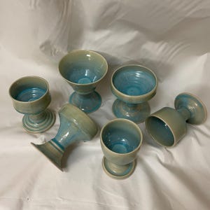 May include: Six handmade ceramic goblets with a turquoise and pale green glaze. The goblets have a rounded bowl shape and are set on a pedestal base. The glaze has a glossy finish, and the goblets are arranged on a white surface.