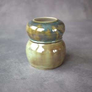 May include: A handmade ceramic jar with a two-part design. The top section is a rounded shape with a teal and brown glaze, while the bottom section is a similar shape with a lighter green and brown glaze. The jar has a glossy finish.