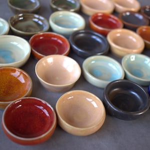 May include: A collection of small, handmade ceramic bowls in various colors, including red, brown, black, and beige. The bowls have a glossy finish and are arranged on a gray surface. Some bowls have a light blue interior.
