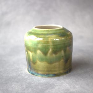 May include: A small, handmade ceramic vase with a cylindrical shape. The vase has a glossy, layered glaze in shades of green and yellow, with a hint of blue at the base. The interior is a light cream colour.