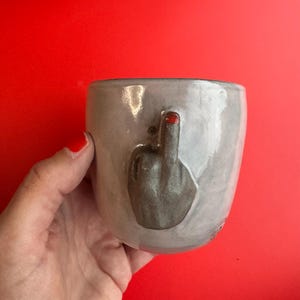 Könnte beinhalten: Ein hellgrauer Keramikbecher mit einer dreidimensionalen, skulptierten Hand auf der Außenseite. Die Hand zeigt eine Mittelfinger-Geste, wobei die Spitze des erhobenen Fingers in leuchtendem Rot bemalt ist und einen Kontrast zum grauen Becher bildet.