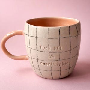 Könnte beinhalten: Ein Keramikbecher mit hellrosa Glasur und einem handgezeichneten dunklen Gittermuster. Der Becher zeigt den eingeprägten Text "fuck off. I'm overthinking" auf der Vorderseite. Er hat einen geschwungenen Henkel und eine glatte, runde Form.