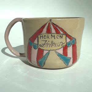 Könnte beinhalten: Eine beige Keramiktasse mit einem rosa Griff. Die Tasse zeigt ein handgezeichnetes Zirkuszelt-Design in Rot, Rosa und Blau mit dem Text "Hormon Zirkus" auf einem gelben Banner. Ein einzigartiges, skurriles Design.