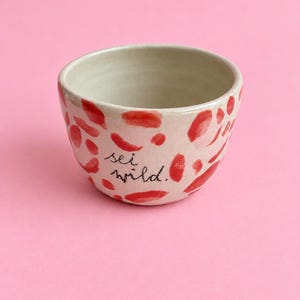 Könnte beinhalten: Eine kleine, cremeweiße Keramikschale mit einem handgemalten Muster aus unregelmäßigen roten Flecken. Die Außenfläche zeigt den schwarzen, handschriftlichen Text "sei wild." in Schreibschrift zwischen den roten Farbakzenten.