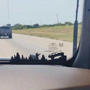 May include: Black silhouette car window decal depicting a mountain scene with trees, a person, and a 4x4 vehicle. The decal is mounted on a car dashboard with a highway and another vehicle visible through the windscreen.