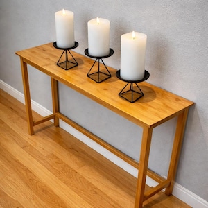 Puede incluir: Una consola de madera con tres velas de pilar blancas en candelabros geométricos negros. Las velas están encendidas, proyectando un brillo cálido. La mesa es de madera clara y está colocada contra una pared gris.