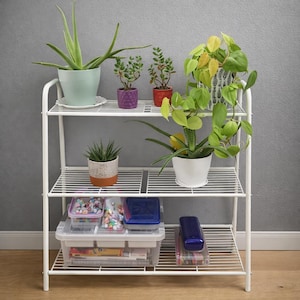 May include: A white metal shelving unit with three tiers, displaying various potted plants and storage containers. The plants include aloe vera, succulents, and trailing vines. Clear plastic containers hold craft supplies and other items. The unit is against a grey wall.