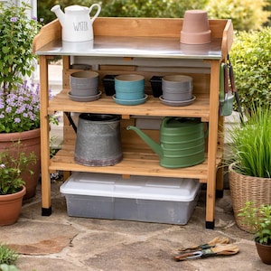 May include: A wooden potting bench with a stainless steel top, holding a white watering can with "WATER" printed on it, terracotta pots, and various planters. The bench has multiple shelves with gardening tools and a green watering can.