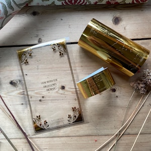 May include: A clear rectangular card with gold trim and the text "STAY WITH THE HIGHLORD, HUMAN" and "EP EDITION". A gold cylinder and a gold band are also present. Floral patterned rolls of paper are in the background.