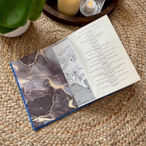 Pode incluir: Um livro de capa dura aberto com um design de mármore cinza escuro e dourado no lado esquerdo e um mapa impresso na aba interna. A página direita exibe uma lista de elogios críticos intitulada Praise for Antonia Hodgson.