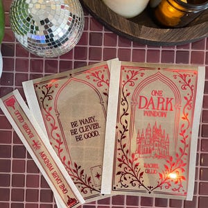 May include: Two translucent vellum overlays and a bookmark featuring red foil text and illustrations. One overlay displays the title One Dark Window with a castle graphic, while the other reads Be Wary. Be Clever. Be Good. The bookmark includes the author name Rachel Gillig.