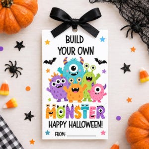 May include: A rectangular white gift tag featuring a black satin ribbon bow at the top. The text reads BUILD YOUR OWN MONSTER HAPPY HALLOWEEN. It displays colorful cartoon monsters in blue, purple, orange, green, and pink with various eyes.