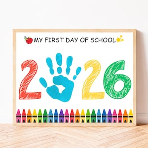 Può includere: Una stampa incorniciata con il testo MY FIRST DAY OF SCHOOL, una mela rossa e stelle gialle. Il numero 2026 è visualizzato con un'impronta di mano blu che sostituisce lo zero. Una fila di pastelli colorati borda la parte inferiore dello sfondo bianco.