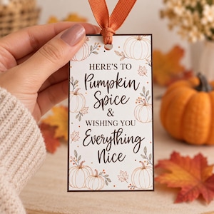 Op de afbeelding: Een rechthoekig wit cadeaulabel met een donkerbruine rand en een oranje lintlus aan de bovenkant. Het label bevat de tekst "HERE'S TO Pumpkin Spice & WISHING YOU Everything Nice", omringd door illustraties van oranje pompoenen en herfstbladeren.