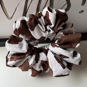 May include: A large scrunchie with a cow print design. The fabric is white with brown spots. The scrunchie is gathered and tied in a bow shape. The background is a white surface with a black and white design.