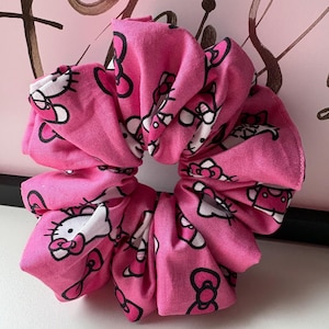 May include: A pink scrunchie featuring a repeating pattern of the Hello Kitty character in white and black. The fabric is gathered and ruched, creating a voluminous, circular shape. The scrunchie is designed for use in hair styling.