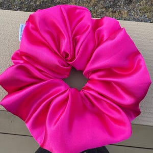 May include: A large, vibrant fuchsia satin scrunchie. The scrunchie is made of a soft, shiny fabric and is gathered in the center. The scrunchie is a hair accessory.