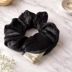 May include: Black scrunchie with a hidden zipper pocket, holding a twenty-dollar bill. The scrunchie is made of a soft, velvety material. The background features gold rings and white flowers.