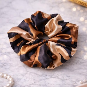 Peut inclure: Un grand chouchou à motifs noirs, marron et dorés. Le tissu satiné est froncé et torsadé, créant un accessoire volumineux. Cet élastique à cheveux est un ajout élégant à toute coiffure.