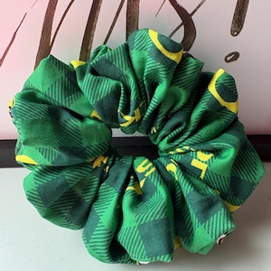 Può includere: Un elastico per capelli a quadri verdi e gialli. Il tessuto è verde brillante con un motivo a quadri gialli. L'elastico è arricciato e ha un elastico nero.