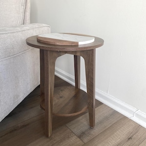 May include: A small, round wooden side table with a lower shelf and four legs. A decorative serving board with a white marble surface and a wooden edge rests on top. The table is approximately 24 inches tall.