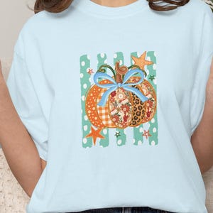 May include: A light blue t-shirt featuring a central graphic of a pumpkin with a light blue bow. The pumpkin has segments with orange gingham, floral, and leopard print patterns. The background consists of teal vertical stripes with white dots.