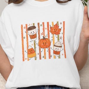 Peut inclure: Un t-shirt blanc avec une impression graphique représentant une tasse de café, une feuille d'érable, une citrouille, un bonbon maïs et une autre tasse de café avec de la crème fouettée. Chaque élément a un visage souriant et des membres.