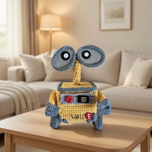 Può includere: Un giocattolo robot all'uncinetto fatto a mano in giallo, grigio e blu, che assomiglia al personaggio Wall-E. Il robot ha grandi occhi grigi, un corpo giallo e il nome "WALL E" è visibile sul davanti. È appoggiato su un tavolo di legno.