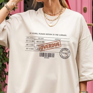 May include: An off-white short-sleeve t-shirt featuring a black graphic of a library checkout card with the text IF FOUND, PLEASE RETURN TO THE LIBRARY, a barcode, and a circular stamp. A red rectangular stamp reads OVERDUE across the center.