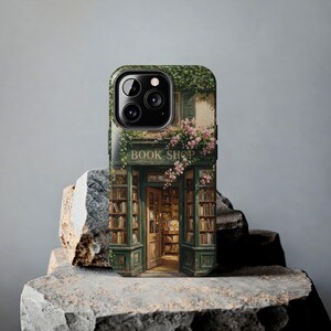 K&ouml;nnte beinhalten: Eine Smartphone-H&uuml;lle mit einer aufgedruckten Illustration eines gr&uuml;nen Ladenlokals und dem Schriftzug BOOK SHOP &uuml;ber der T&uuml;r. Das Design zeigt Regale voller B&uuml;cher, Kletterpflanzen und rosa Blumen rund um den Eingang des Gesch&auml;fts.