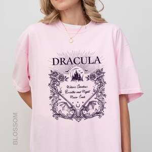 May include: A light pink short-sleeve t-shirt featuring a dark purple graphic print. The design includes the text "DRACULA" and "Where Shadows Breathe and Night Never Ends," surrounded by a decorative frame of roses, bats, and a castle.