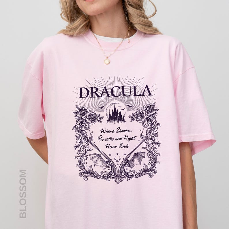 May include: A light pink short-sleeve t-shirt featuring a dark purple graphic print. The design includes the text "DRACULA" and "Where Shadows Breathe and Night Never Ends," surrounded by a decorative frame of roses, bats, and a castle.