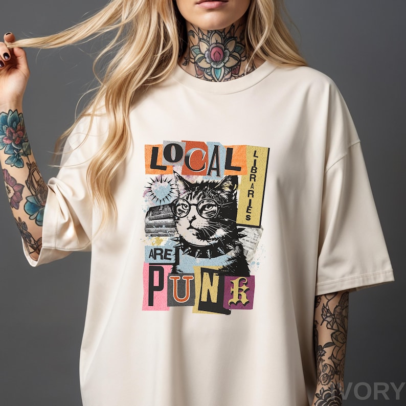 May include: An ivory short-sleeve t-shirt featuring a graphic print of a cat wearing round glasses and a spiked collar. The text reads LOCAL LIBRARIES ARE PUNK, arranged in a collage style with colourful rectangular blocks and abstract shapes.