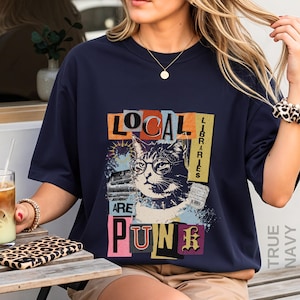 May include: A dark navy blue short-sleeve t-shirt featuring a graphic print of a cat wearing glasses. The text reads "LOCAL LIBRARIES ARE PUNK" in a collage style with various colours and textures. The design includes a stack of books and a receipt.