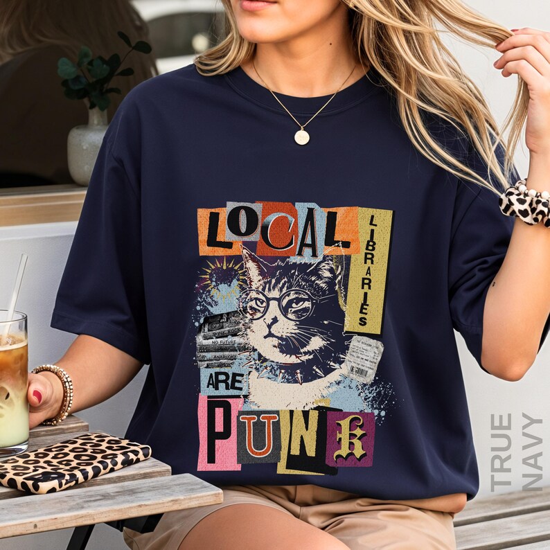 May include: A dark navy blue short-sleeve t-shirt featuring a graphic print of a cat wearing glasses. The text reads "LOCAL LIBRARIES ARE PUNK" in a collage style with various colours and textures. The design includes a stack of books and a receipt.