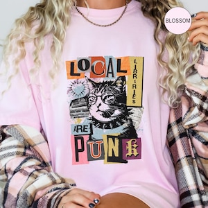May include: A light pink t-shirt featuring a graphic of a cat wearing round glasses and a spiked collar. The text reads "LOCAL LIBRARIES ARE PUNK" in a collage style with various fonts and colours. The design includes a heart and book imagery.