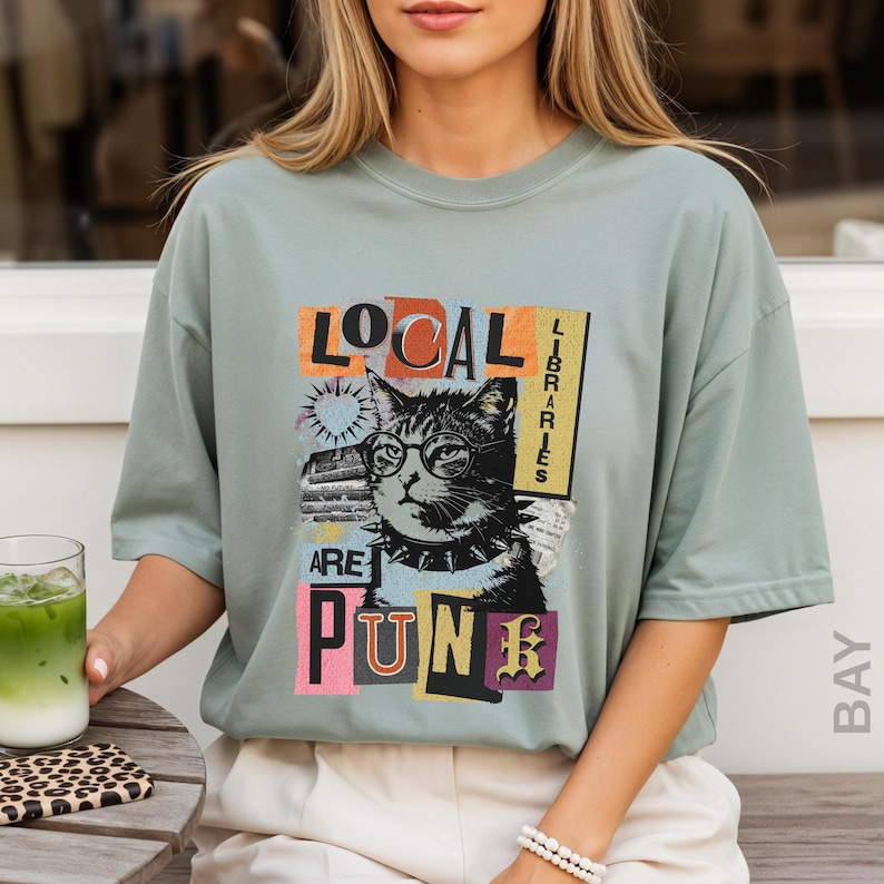 May include: A sage green short-sleeve t-shirt featuring a graphic of a cat wearing glasses and a spiked collar. The text reads "LOCAL LIBRARIES ARE PUNK" in a collage-style font with orange, yellow, and pink accents. The design includes a sunburst icon.